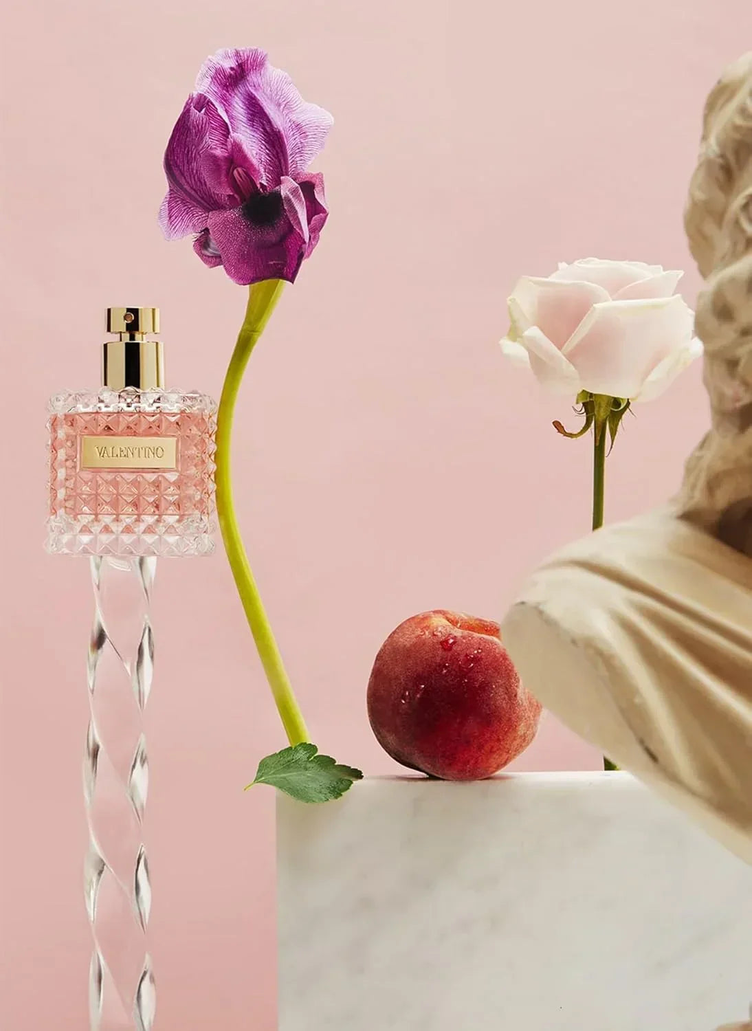 Valentino fragrance bottle with pink crystal design, purple flower, peach, and marble bust on pink background