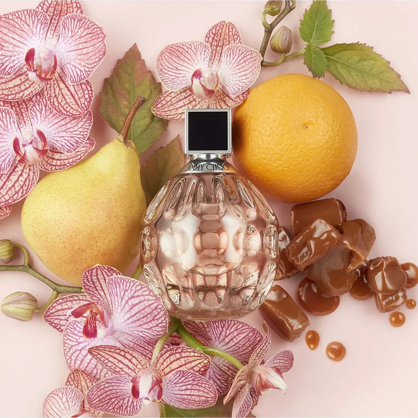 Perfume bottle surrounded by pink orchids, a lemon, a pear, and chocolate pieces on a light background