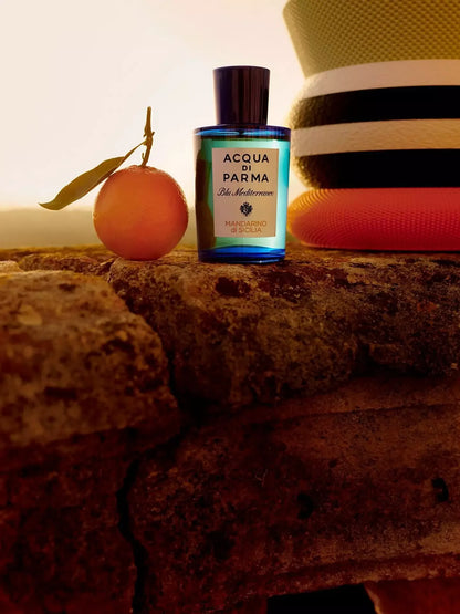 Acqua di Parma Il Mediterraneo cologne bottle with a blue glass body and black cap, styled on a rustic stone surface next to an orange and a striped hat.