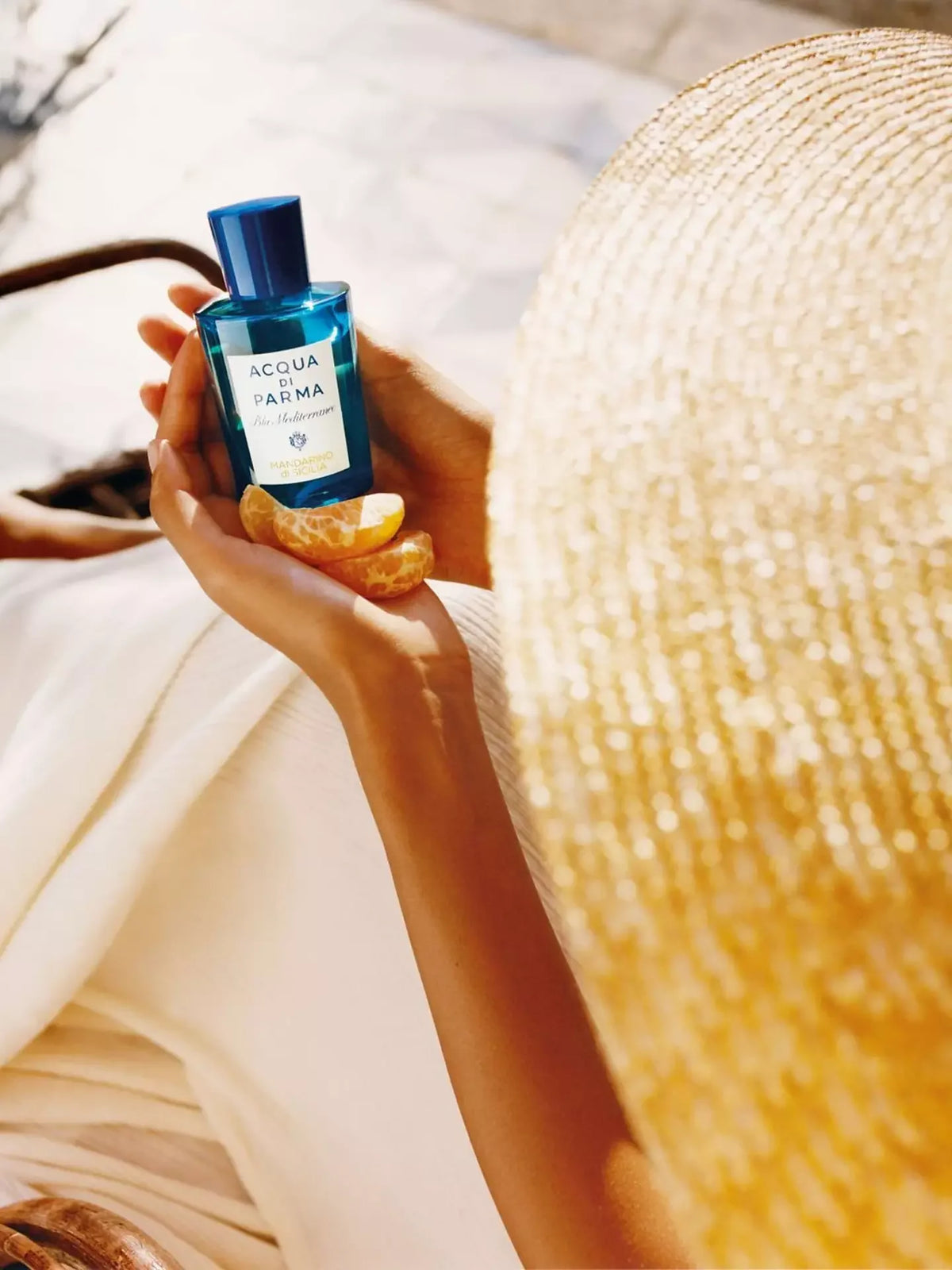 Person holding a blue bottle labeled 'Acqua di Parma' with a straw hat in the background