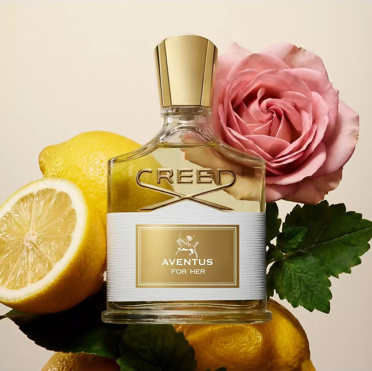 Creed Aventus For Her perfume bottle with a gold cap and white-and-gold label, styled with lemons, a pink rose, and green leaves on a warm beige background.