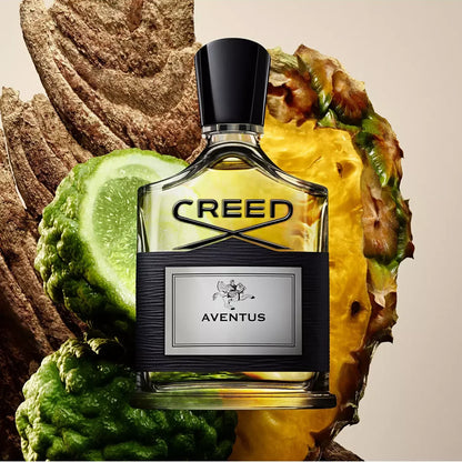 A bottle of Creed Aventus fragrance centered against a styled background of tropical fruits, including a pineapple, guava, and banana, on a light beige background.