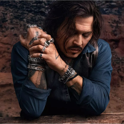 A man in a dark blue shirt rests his head on his arms, wearing multiple silver chain bracelets, rings, and a necklace, set against a dark, rocky background.
