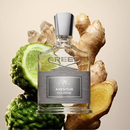 A bottle of Creed Aventus Cologne centered against a warm beige background, surrounded by fresh ingredients including ginger roots, a lime slice, and a bitter melon. The bottle is clear glass with a brushed silver label and a polished silver cap.