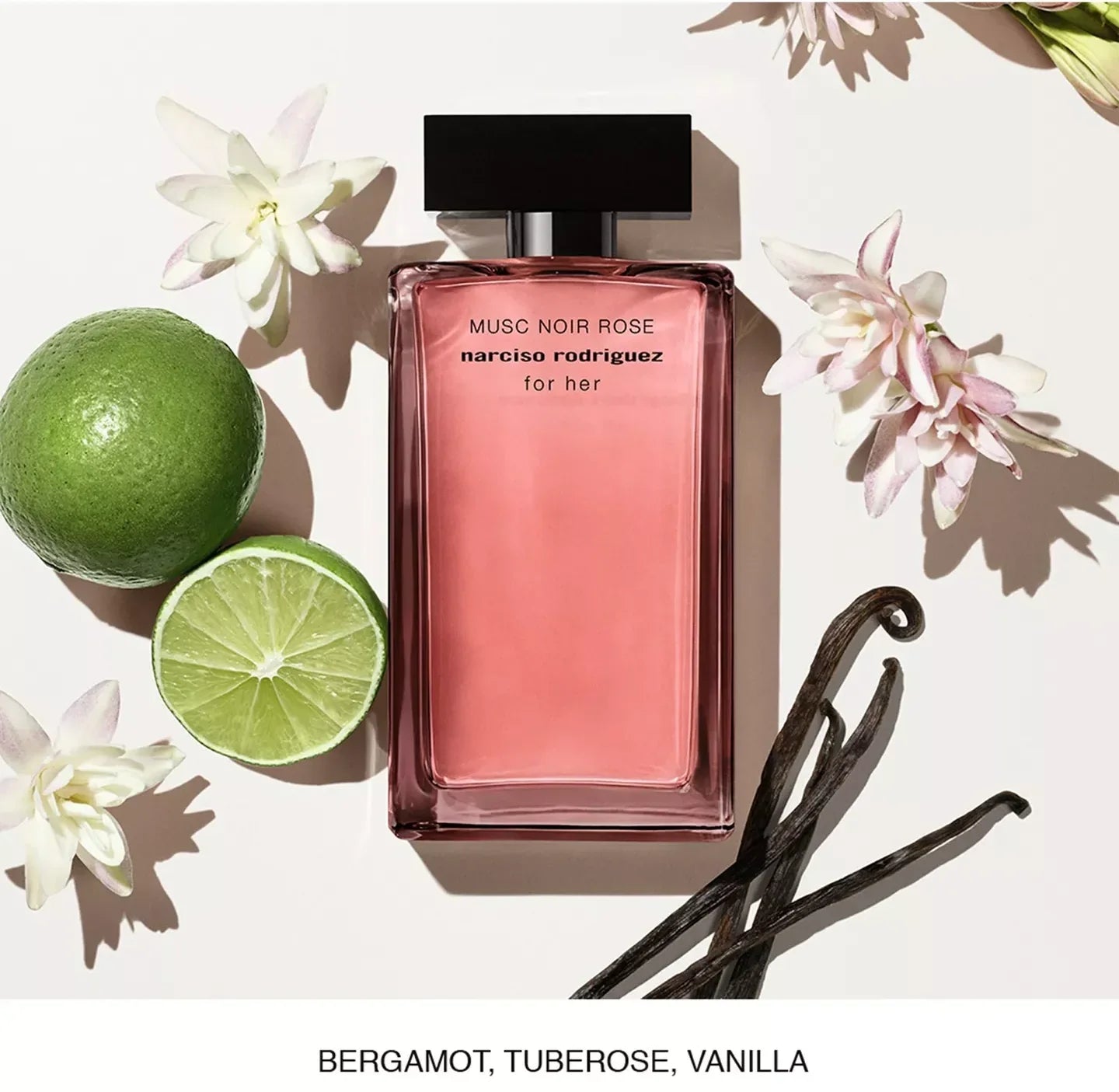 A rectangular pink perfume bottle labeled 'MUSC NOIR ROSE narciso rodriguez for her' is arranged on a white background with a lime, white flowers, vanilla beans, and a text banner at the bottom reading 'BERGAMOT, TUBEROSE, VANILLA'.