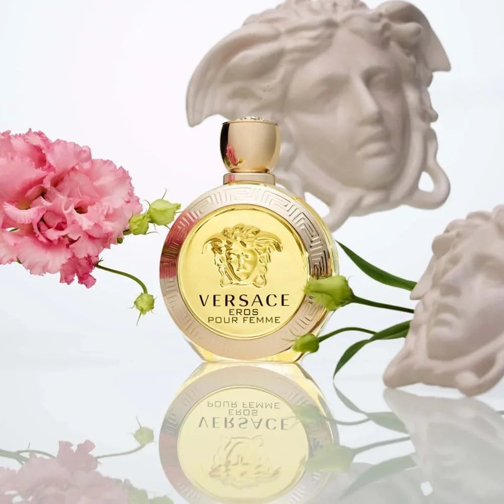 Versace Eros Pour Femme perfume bottle in gold, styled with pink carnations and white classical busts on a reflective white surface.
