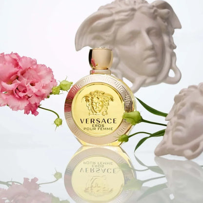 Versace Eros Pour Femme perfume bottle in gold, styled with pink carnations and white classical busts on a reflective white surface.