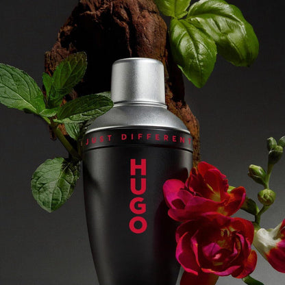Black Hugo perfume bottle with red text, surrounded by green leaves and flowers on a dark background