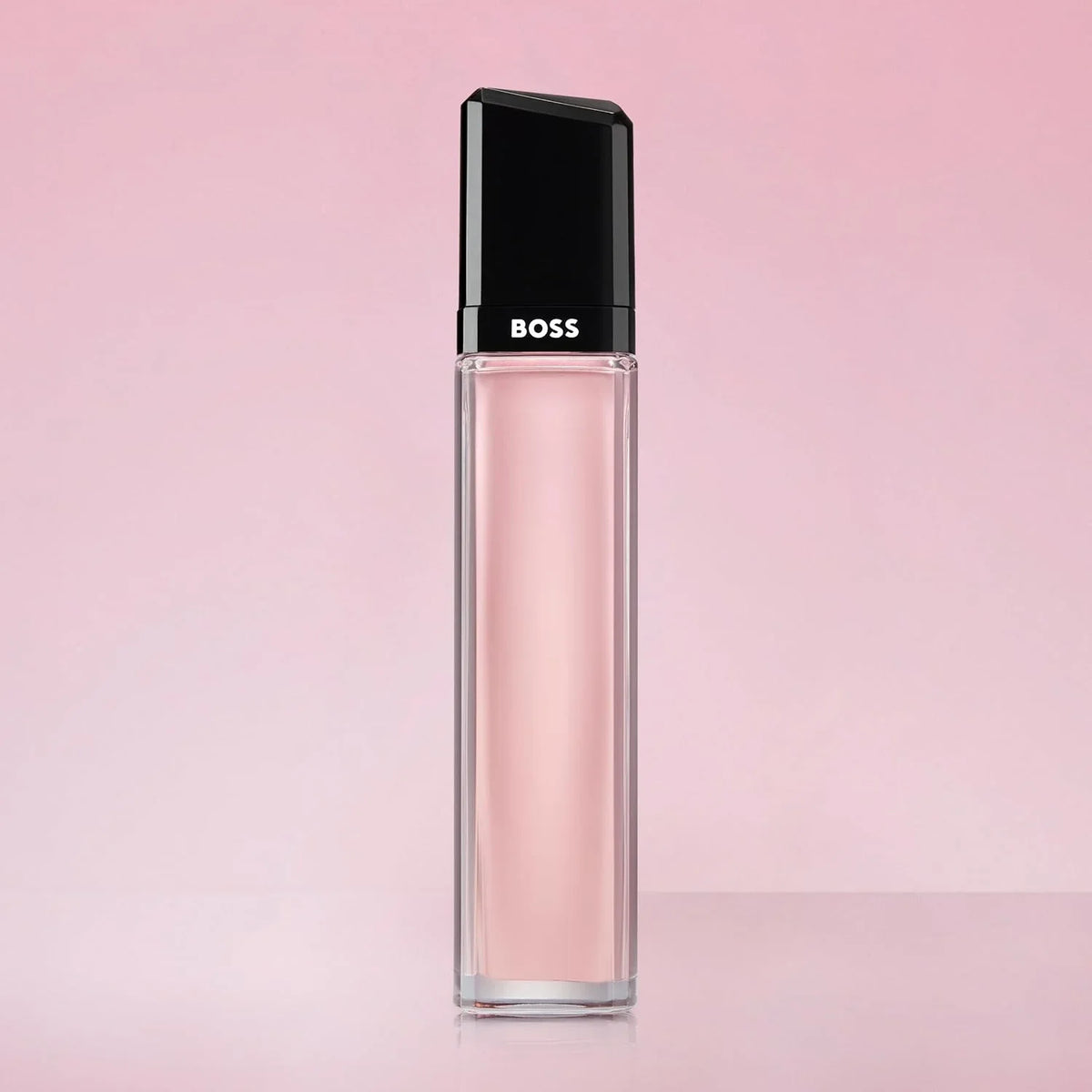 A clear rectangular lip gloss bottle with a black cap, filled with light pink gloss, standing on a matching pink surface against a soft pink background. The cap is printed with the white BOSS logo.
