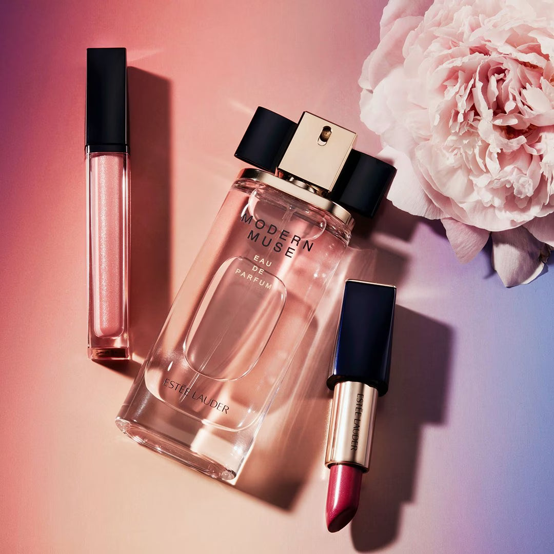 A styled flat-lay of three cosmetic products on a pink-to-purple gradient background with a large pale pink flower: a clear rectangular perfume bottle labeled 'MODERN MUSE EAU PARFUMEE' by Stéphane Leroux, a pink lip gloss tube, and a pink lipstick with a black cap and gold base.