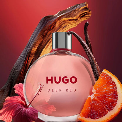 Hugo Deep Red perfume bottle with a pink liquid, styled with a hibiscus flower, a slice of blood orange, and a vanilla bean against a red gradient background.