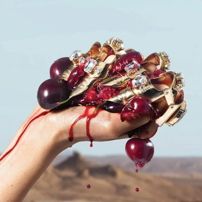 Hand holding crushed cherries and gold rings with gemstones, juice dripping against desert backdrop