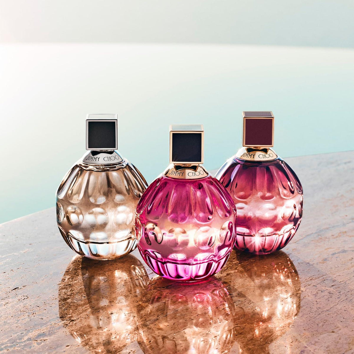 Three sculptural perfume bottles with faceted glass and square caps are arranged on a reflective stone surface against a soft pale blue background. The bottles are translucent champagne beige, pink, and magenta, with gold-toned detailing and dark square accents on the caps. 60ml, 100ml
