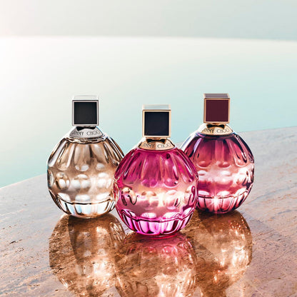Three sculptural perfume bottles with faceted glass and square caps are arranged on a reflective stone surface against a soft pale blue background. The bottles are translucent champagne beige, pink, and magenta, with gold-toned detailing and dark square accents on the caps. 60ml, 100ml