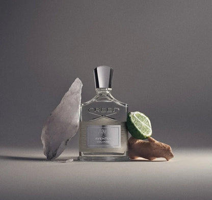 A bottle of Creed Aventus cologne stands on a gray studio surface, flanked by a large translucent crystal on the left and a sliced lime and piece of ginger on the right.