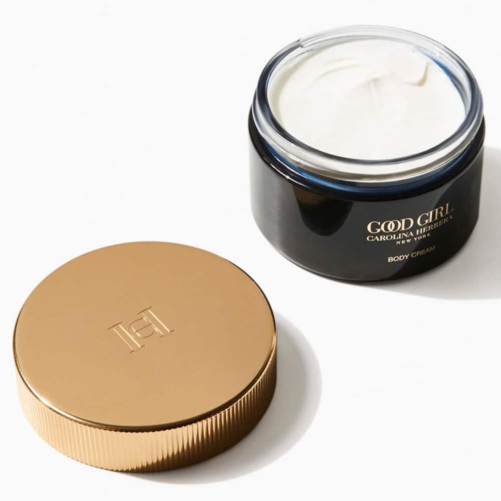 A jar of Carolina Herrera Good Girl Body Cream shown open with the white cream visible, alongside its metallic gold screw-on lid on a clean white background.