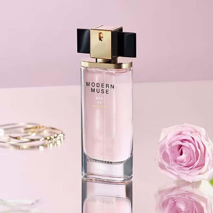 Estée Lauder Modern Muse Eau de Parfum bottle with a gold and black cap, set on a reflective pink surface with a pink rose and gold jewelry in the background.