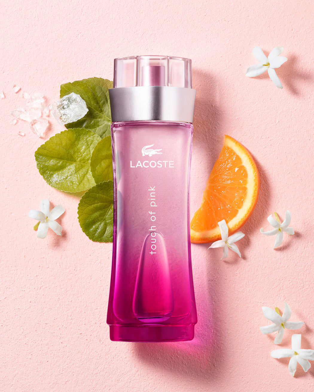 Pink Lacoste perfume bottle on a pink background with flowers and an orange slice.