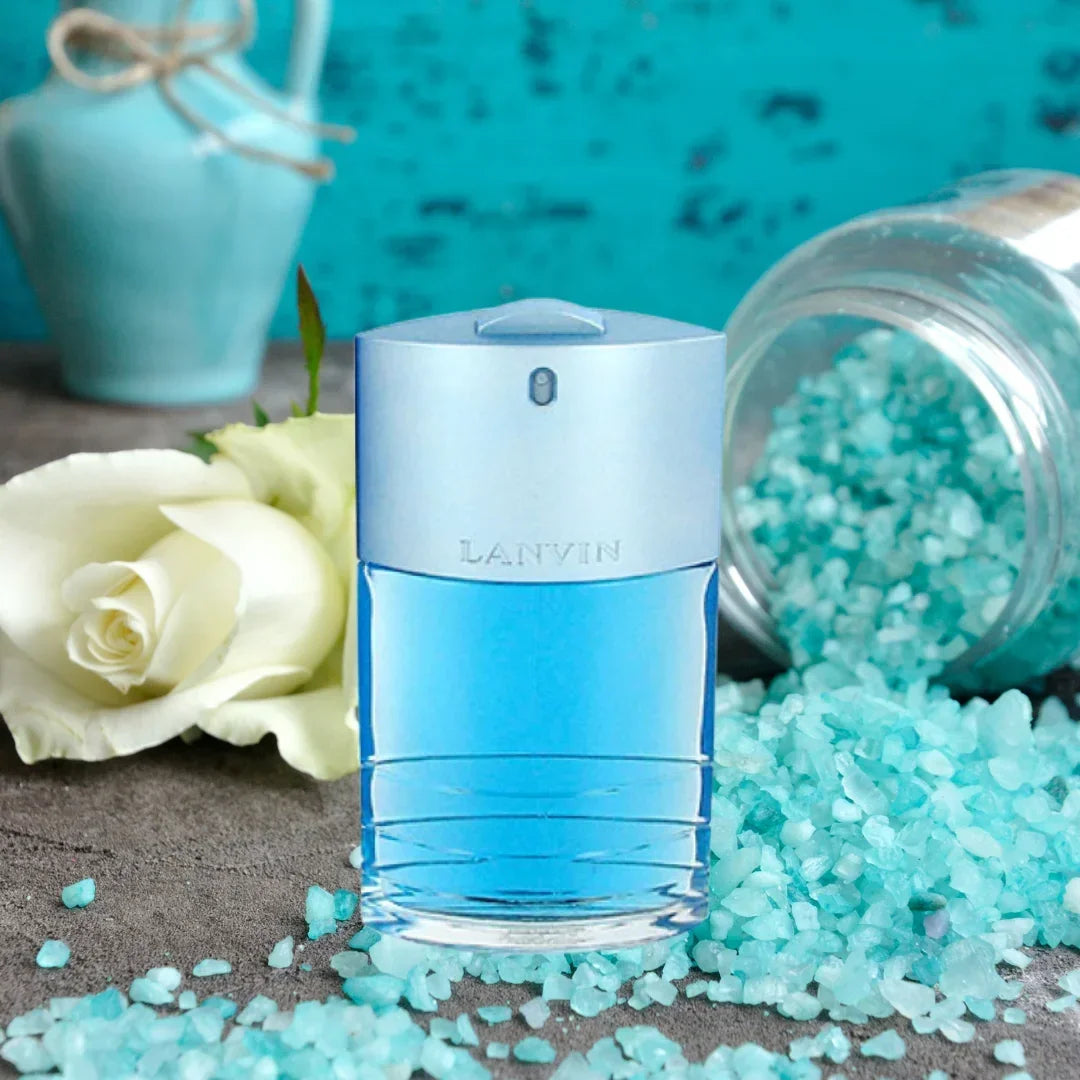 A bottle of Lanvin Blue fragrance centered on a textured surface, surrounded by light blue salt crystals, a white rose, and a turquoise decorative vase in the background.