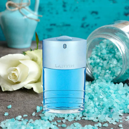A bottle of Lanvin Blue fragrance centered on a textured surface, surrounded by light blue salt crystals, a white rose, and a turquoise decorative vase in the background.