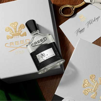 Creed Aventus perfume bottle on a wooden surface with a box and card in the background.